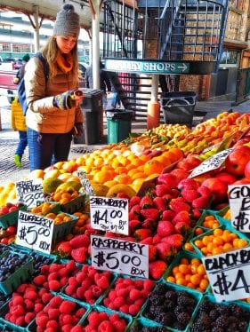 Chai está segurando a metade de uma laranja atrás de uma banca de frutas vermelhas no Pike Place Market de Seattle