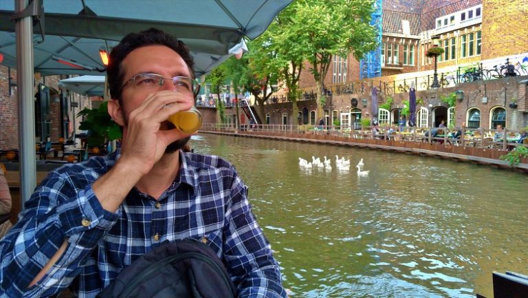 Augusto de camisa xadrez está bebendo uma cerveja e ao fundo um canal onde um grupo de cisne nada tranquilamente