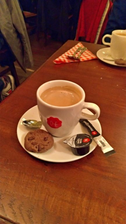 Na foto uma xícara branca de café expresso com um biscoito, sachê de açúcar e potinho de leite no pires sobre a mesa de madeira. Belgisch Biercafé Olivier Utrecht