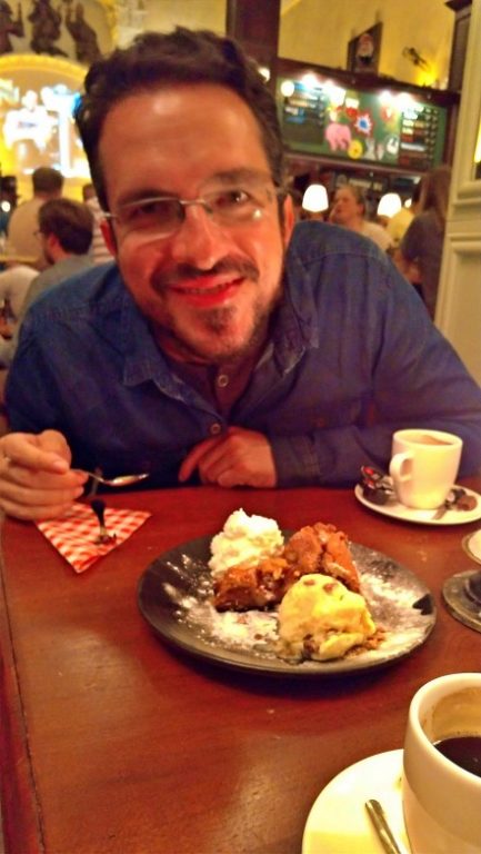 Na foto Augusto está sorrindo com a colher na mão aguardando para comer a fatia de torta de maça entre uma bola de sorvete e uma porção de chantili que está no prato a sua frente