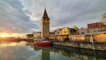Sol se pondo no canto inferior esquerdo refletindo na água do pier onde no centro da foto se vê a torre alta e antiga de telhados coloridos, um barco pequeno ancorado e os demais prédios do porto