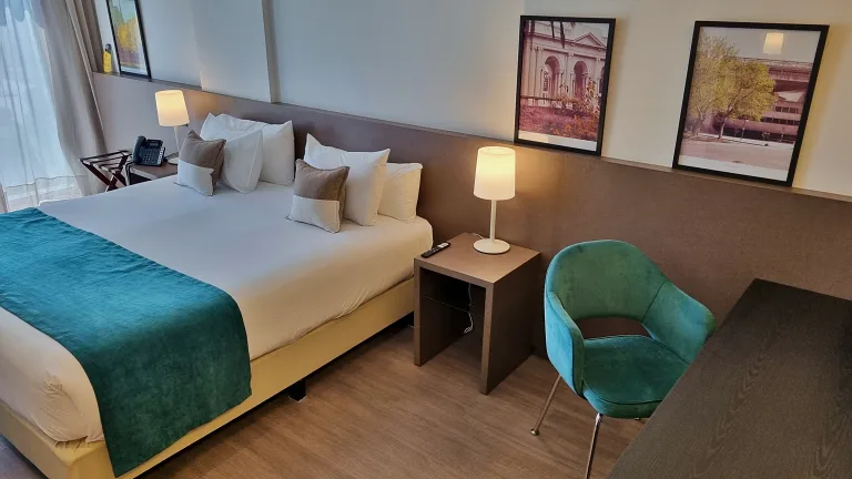 Onde ficar em La Plata: Conheça Dazzler by Wyndham La Plata 13 Cama king size no quarto da suíte do Dazzler by Wyndham La Plata, com decoração moderna, travesseiros confortáveis e uma cadeira verde ao lado da mesa
